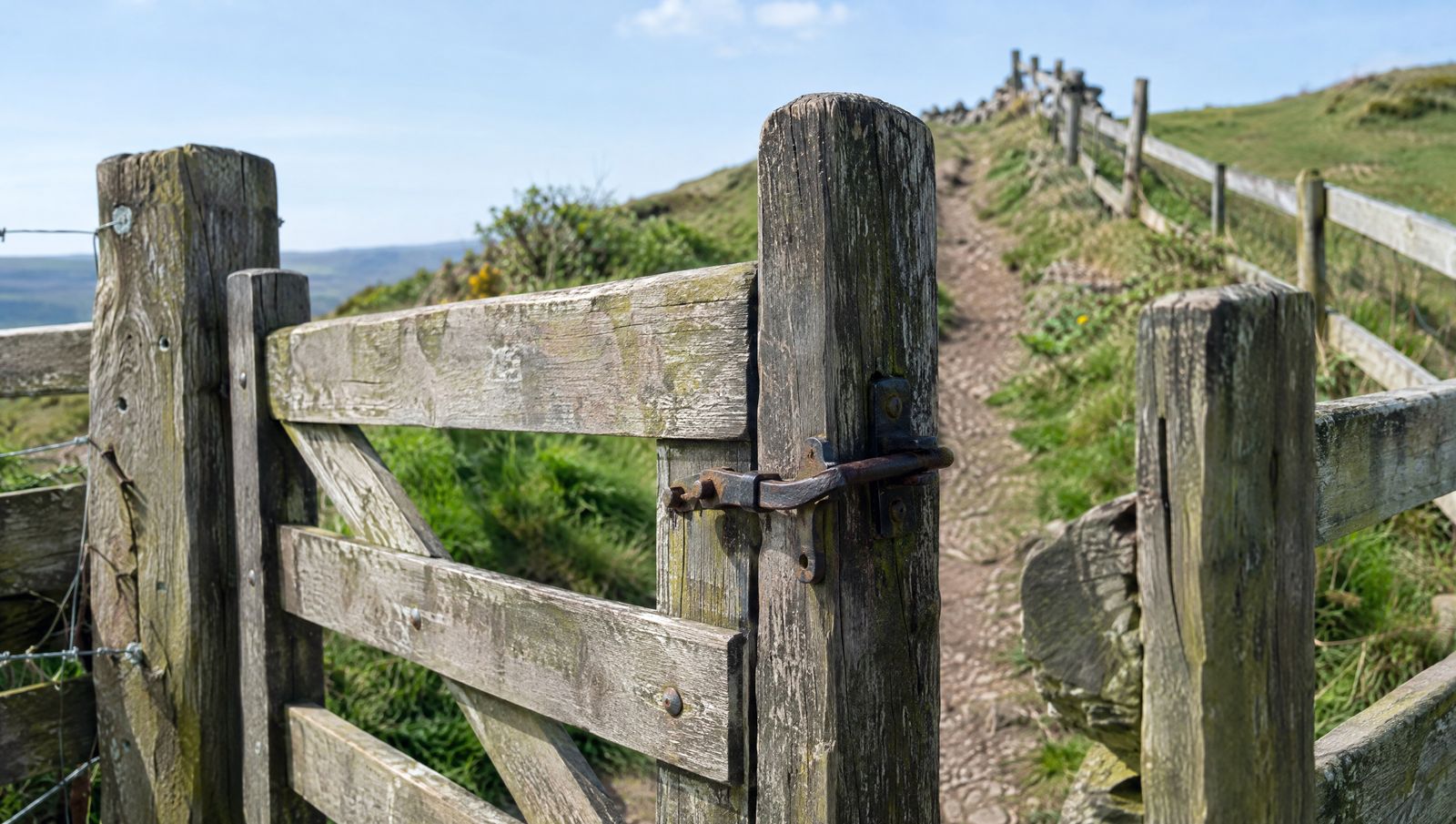 A weathered wooden kissing gate with an iron latch on a hillside footpath